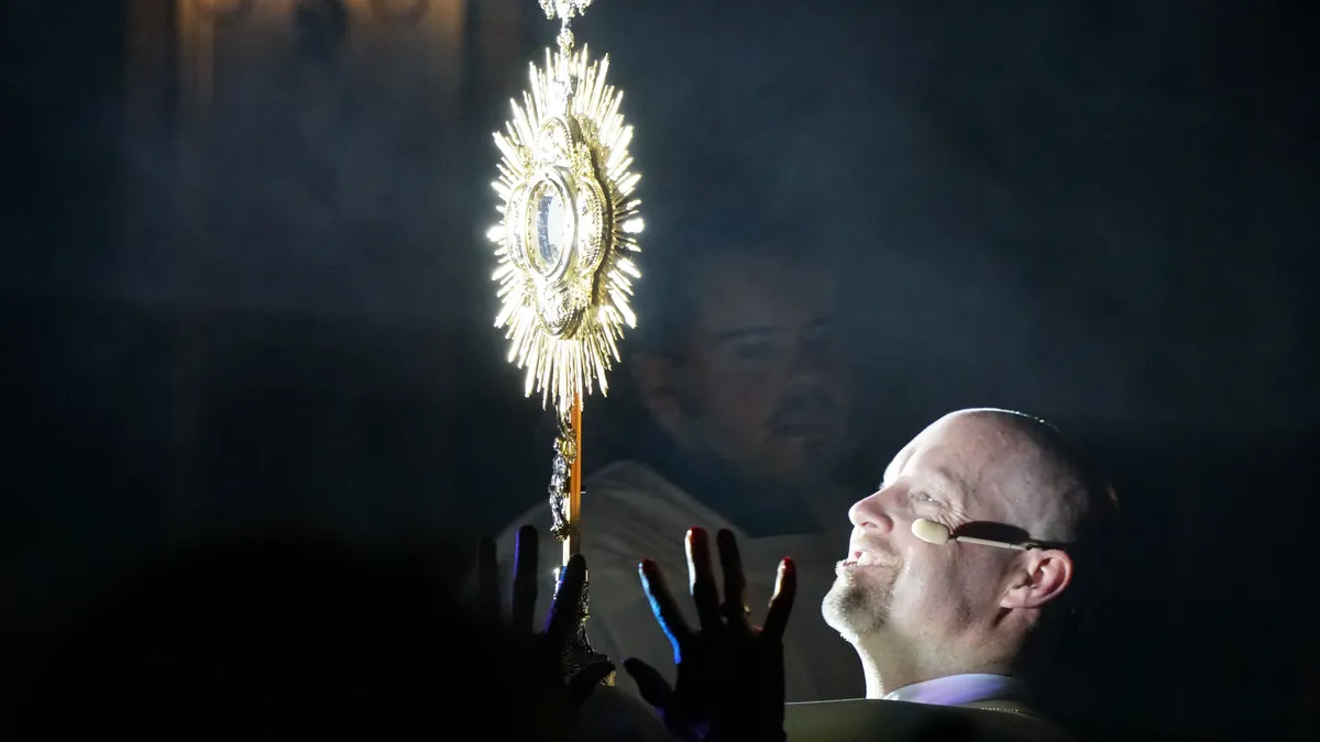A priest holds a gold monstrance amid incense and raised hands