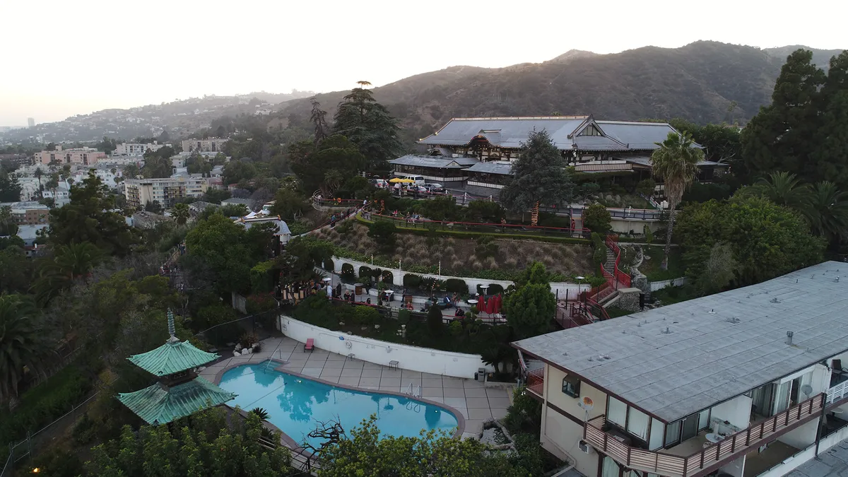 An aerial view of buildings, a pool, and wooded hills in evening light
