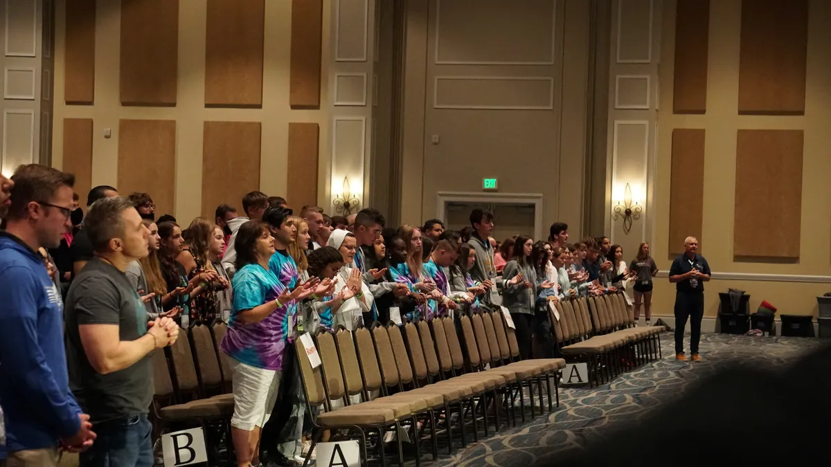 Conference attendees stand in rows of chairs inside a large hall