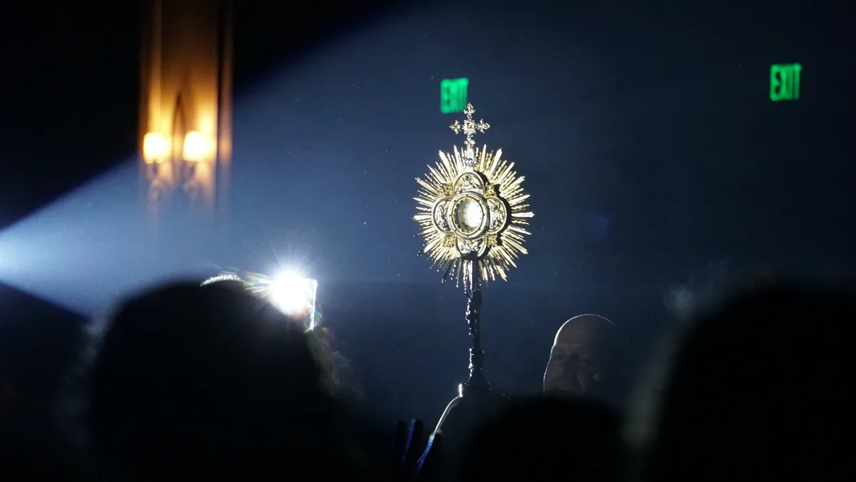 An illuminated monstrance seen above silhouettes of the congregation